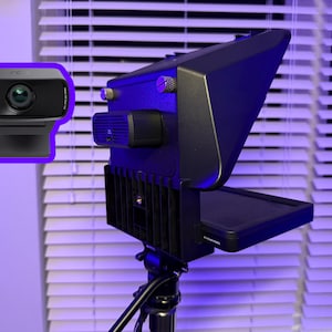 May include: A black webcam with a built-in microphone and a black video teleprompter. The teleprompter is mounted on a tripod. The background includes a window with white blinds.