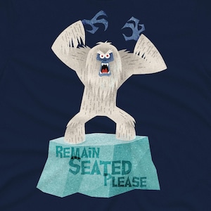 Puede incluir: Una camiseta azul marino con una ilustración de un monstruo yeti blanco. El yeti está de pie sobre un bloque de hielo azul con el texto "REMAIN SEATED PLEASE".