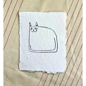May include: A simple black and white illustration of a cat. The cat is depicted with basic lines, sitting on textured white paper. The paper has a rough, torn edge. The background is a beige fabric with red stripes.