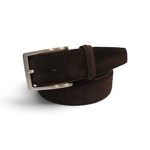 May include: A dark brown suede belt featuring a rectangular silver-toned metal buckle and a matching suede belt loop. The belt is coiled in a circular shape, showcasing the textured material and the metallic hardware finish clearly.