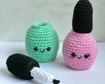 May include: Two crocheted nail polish bottles, one green and one pink, with cute faces. The green bottle is open and the pink bottle has a black cap. A black and white crochet brush is lying on the table.