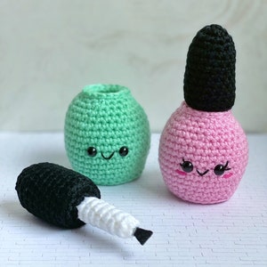 Puede incluir: Dos botellas de esmalte de uñas de crochet, una verde y otra rosa, con caras lindas. La botella verde está abierta y la botella rosa tiene una tapa negra. Un pincel de crochet negro y blanco está sobre la mesa.
