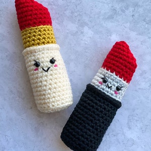 Puede incluir: Dos juguetes de labios de crochet con caras sonrientes. Uno es blanco con una banda amarilla y una parte superior roja, el otro es negro con una parte superior roja.