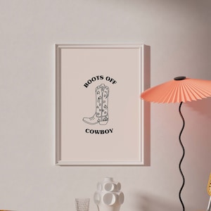 May include: A framed print with the text "Boots Off Cowboy" and a black and white illustration of a cowboy boot. Two wicker chairs with metal frames and a small wooden table with a white ceramic vase and a glass on top are in the background.