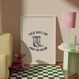 May include: A framed print with the text "These boots are made for walkin'" and a black and white illustration of two cowboy boots. The print is hanging on a light beige wall above a green and white checkered rug. A pink lamp with a white base is on a light green side table.