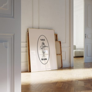 May include: A framed art print featuring a black and white illustration of a person playing a guitar, with the words "Yee Haw" in an oval. The artwork leans against a wall in a room with hardwood floors.