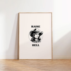 May include: Framed art print featuring a black and white illustration of a woman wearing a cowboy hat. The text "RAISE HELL" is printed above and below the illustration. The artwork is displayed on a white wall above a wooden floor.