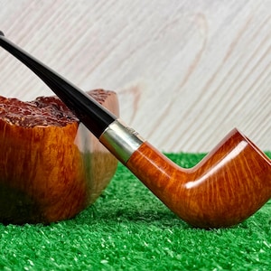 Puede incluir: Una pipa de tabaco con un hornillo de madera de color marr&oacute;n claro liso con veta visible, una boquilla negra pulida y una banda de metal de color plateado que conecta ambos componentes. La pipa est&aacute; colocada en &aacute;ngulo con la boquilla hacia arriba.