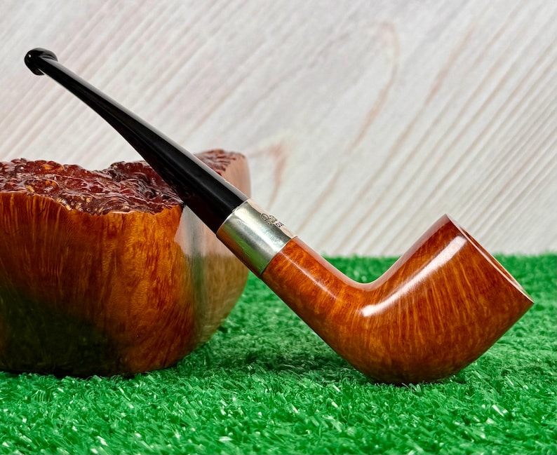 Puede incluir: Una pipa de tabaco con un hornillo de madera de color marr&oacute;n claro liso con veta visible, una boquilla negra pulida y una banda de metal de color plateado que conecta ambos componentes. La pipa est&aacute; colocada en &aacute;ngulo con la boquilla hacia arriba.