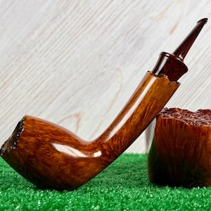 Pode incluir: Um cachimbo de tabaco curvo com um acabamento castanho polido. O cachimbo tem um bocal castanho-avermelhado escuro e uma fornilho texturizado. Está colocado sobre relva artificial verde, com um fundo claro com grão de madeira. O cachimbo tem cerca de 15 cm de comprimento.
