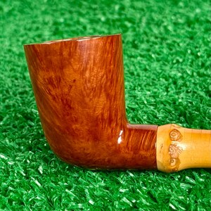 May include: A handcrafted tobacco pipe with a curved, brown wooden bowl and a light-colored stem. The bowl has a rich, swirling grain pattern. The stem is attached with a light-colored band with carved details. The pipe rests on a green surface.
