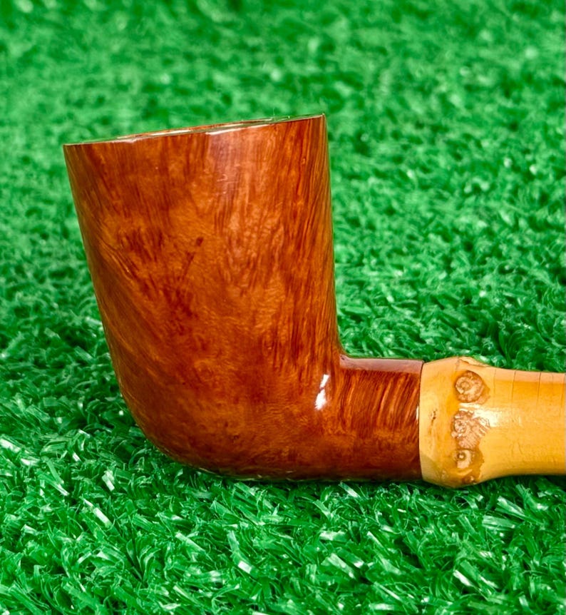 May include: A handcrafted tobacco pipe with a curved, brown wooden bowl and a light-colored stem. The bowl has a rich, swirling grain pattern. The stem is attached with a light-colored band with carved details. The pipe rests on a green surface.