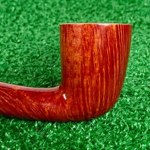 May include: A tobacco pipe crafted from polished wood with a rich, reddish-brown finish. The bowl features a prominent vertical grain pattern that transitions into a smooth, curved shank. The surface has a glossy sheen, highlighting the natural wood texture.
