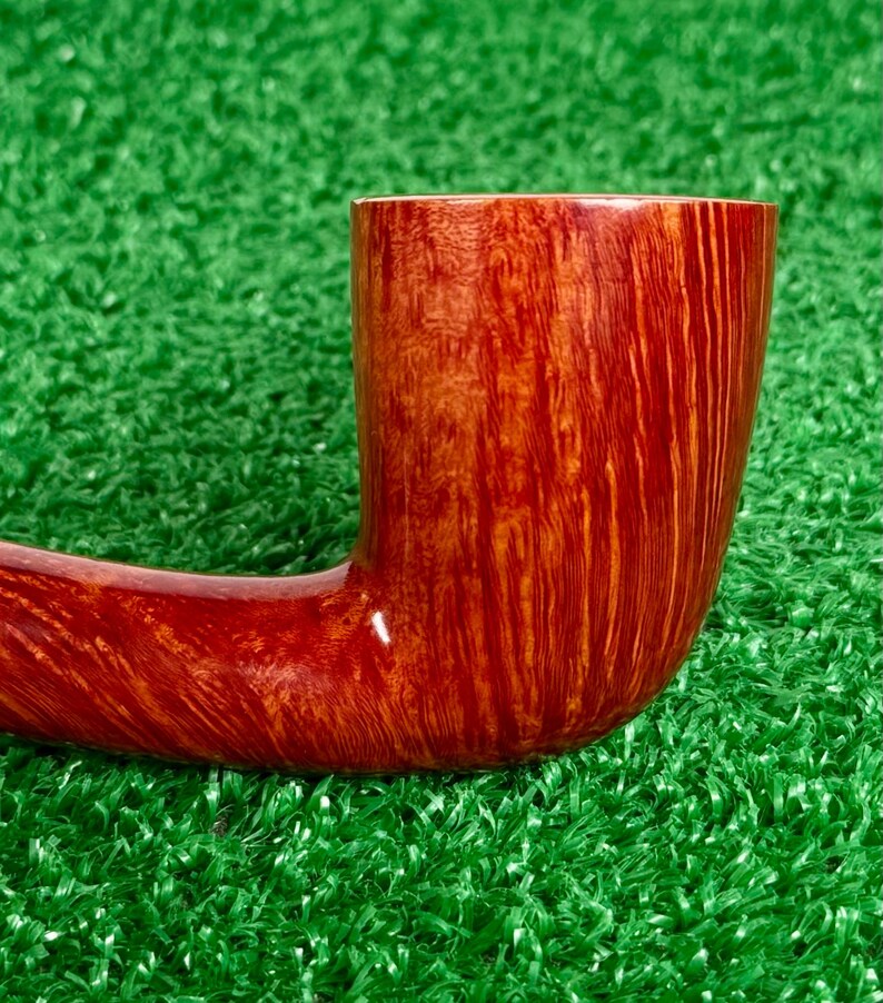 May include: A tobacco pipe crafted from polished wood with a rich, reddish-brown finish. The bowl features a prominent vertical grain pattern that transitions into a smooth, curved shank. The surface has a glossy sheen, highlighting the natural wood texture.