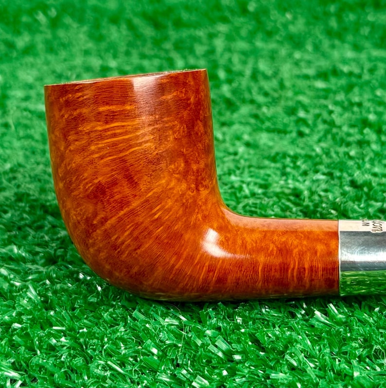Puede incluir: Una pipa de tabaco de madera marr&oacute;n pulida con un acabado de grano natural suave y una banda de metal de color plateado en la base de la boquilla. La pipa presenta un hornillo curvo y un ca&ntilde;o cil&iacute;ndrico con una textura de madera clara.