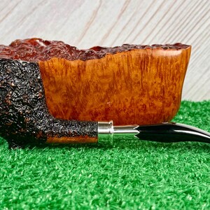 May include: A handcrafted tobacco pipe with a dark, textured bowl and a smooth, light brown wooden body. The stem is black with a silver band. The pipe rests on a green surface.