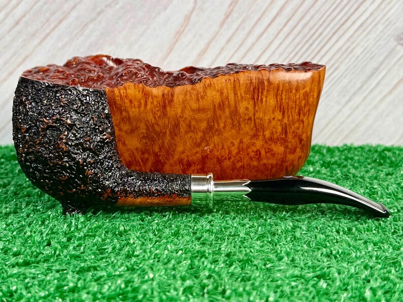 May include: A handcrafted tobacco pipe with a dark, textured bowl and a smooth, light brown wooden body. The stem is black with a silver band. The pipe rests on a green surface.