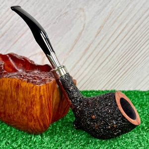 May include: A tobacco pipe with a black stem and a textured, dark brown bowl. The pipe rests on a wooden stand with a rough, natural finish. The pipe has a silver band and a small blue accent.