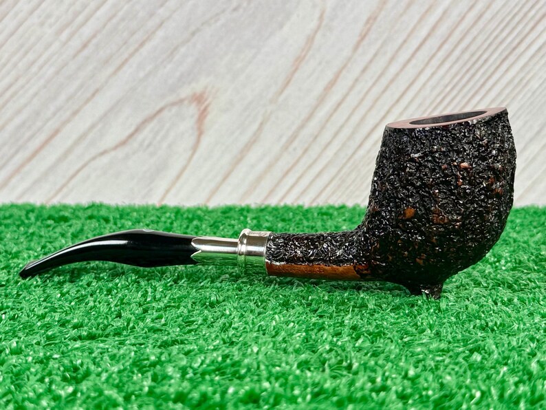 May include: A tobacco pipe with a dark, textured bowl and a curved, black stem. The pipe features a silver band and rests on a green surface. The pipe is designed for smoking tobacco.