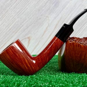May include: A tobacco pipe with a polished reddish-brown wooden bowl featuring a prominent grain pattern. The pipe has a smooth, black acrylic stem that connects to the bowl. The bowl is angled upward, and the stem extends horizontally.