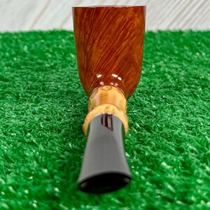 May include: A close-up of a tobacco pipe. The pipe features a brown, polished bowl with a wood grain pattern. The stem is black and the base is a light tan color. The pipe is set against a green background.