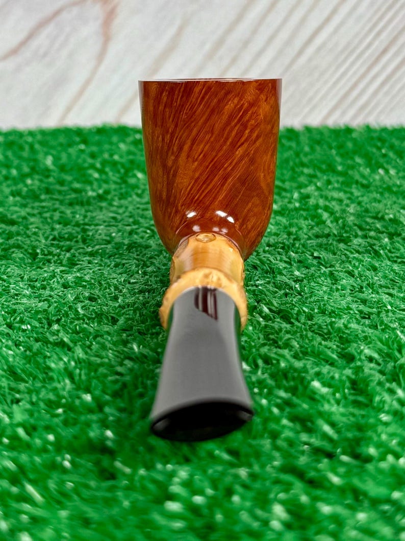 May include: A close-up of a tobacco pipe. The pipe features a brown, polished bowl with a wood grain pattern. The stem is black and the base is a light tan color. The pipe is set against a green background.