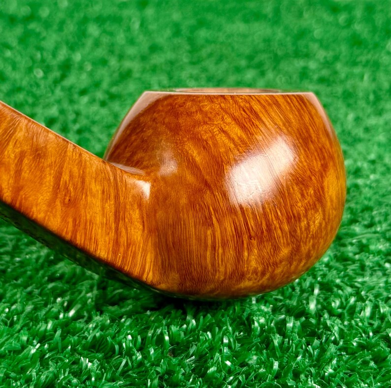 May include: A polished tobacco pipe bowl crafted from light brown wood with a prominent, swirling grain pattern. The smooth, rounded surface reflects light, highlighting the natural texture and warm golden-brown tones of the polished wooden material.