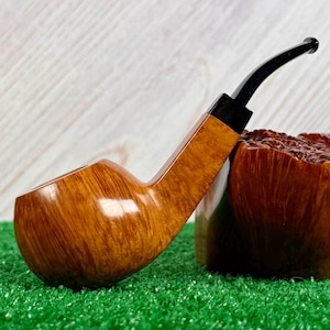 May include: A tobacco pipe with a polished light brown wooden bowl featuring a visible grain pattern and a smooth, curved black stem. The pipe is positioned next to a textured, dark brown wooden block, showcasing a contrast in wood finishes.