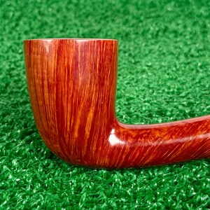May include: A polished tobacco pipe featuring a smooth, reddish-brown wood grain finish with vertical streaks. The bowl is cylindrical and transitions into a curved stem, showcasing a glossy, reflective surface throughout the entire object.