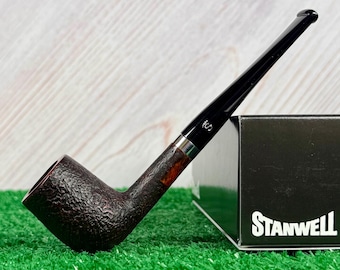 May include: A dark brown tobacco pipe with a textured bowl and a smooth black stem featuring a silver-colored band and a small white S logo. The pipe rests against a black box with the white text STANWELL printed on the side of the box.
