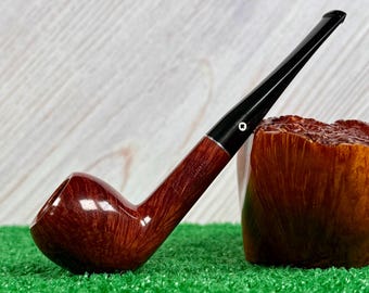 May include: A polished brown wooden tobacco pipe with a smooth, curved bowl and a long, slender black stem. The stem features a small white circular inlay near the shank. The wood grain is visible on the bowl, which has an engraved logo.