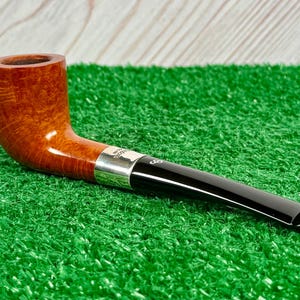 Puede incluir: Una pipa de tabaco con un hornillo de madera marr&oacute;n pulida con veta visible, una banda de metal plateado y una boquilla de acr&iacute;lico negro liso. La boquilla muestra una peque&ntilde;a marca de logotipo blanca cerca de la banda plateada.