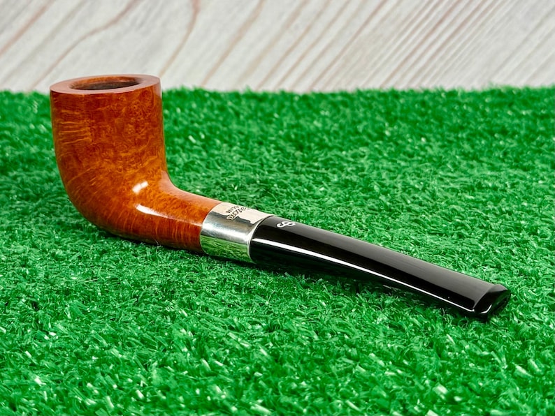 Puede incluir: Una pipa de tabaco con un hornillo de madera marr&oacute;n pulida con veta visible, una banda de metal plateado y una boquilla de acr&iacute;lico negro liso. La boquilla muestra una peque&ntilde;a marca de logotipo blanca cerca de la banda plateada.
