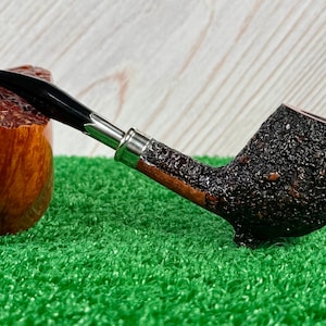 May include: A tobacco pipe with a black stem and a dark, textured bowl. The pipe has a silver band and a wooden stem. The bowl is dark brown and has a rough texture. The pipe is resting on a green surface.
