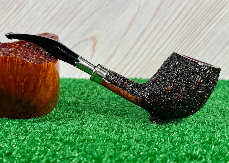 May include: A tobacco pipe with a black stem and a dark, textured bowl. The pipe has a silver band and a wooden stem. The bowl is dark brown and has a rough texture. The pipe is resting on a green surface.