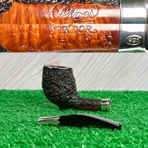 May include: A tobacco pipe with a dark, textured bowl and a curved stem. The pipe has a silver band and a separate black mouthpiece. The words "Pipes", "Srdor", "FRANCE", and "ITALY" are visible on the pipe.