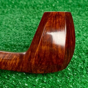 Pode incluir: Um cachimbo de tabaco castanho polido com um acabamento liso e brilhante e um padr&atilde;o de gr&atilde;o de madeira distinto. O cachimbo apresenta um design de fornilho curvo e facetado com uma cor mogno rica e quente e texturas de madeira vis&iacute;veis.
