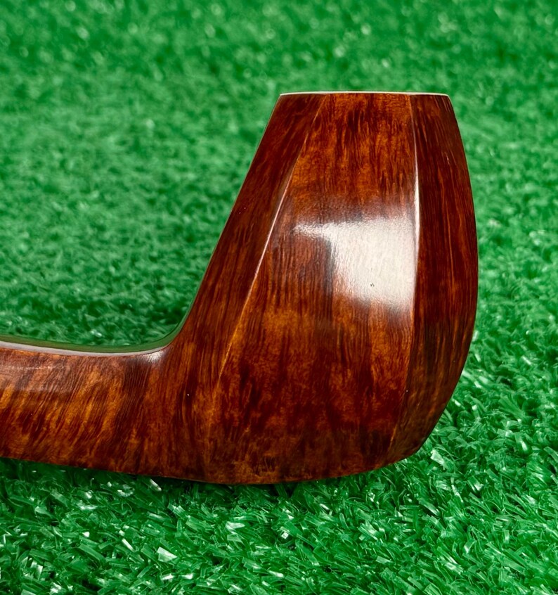 Pode incluir: Um cachimbo de tabaco castanho polido com um acabamento liso e brilhante e um padr&atilde;o de gr&atilde;o de madeira distinto. O cachimbo apresenta um design de fornilho curvo e facetado com uma cor mogno rica e quente e texturas de madeira vis&iacute;veis.