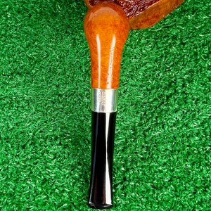 Puede incluir: Una pipa de tabaco con un hornillo de madera de brezo marr&oacute;n pulido con acabado texturizado, unido a un ca&ntilde;o de madera de tono m&aacute;s claro. Una banda de metal plateado separa el ca&ntilde;o de una boquilla c&oacute;nica negra, lisa y brillante.