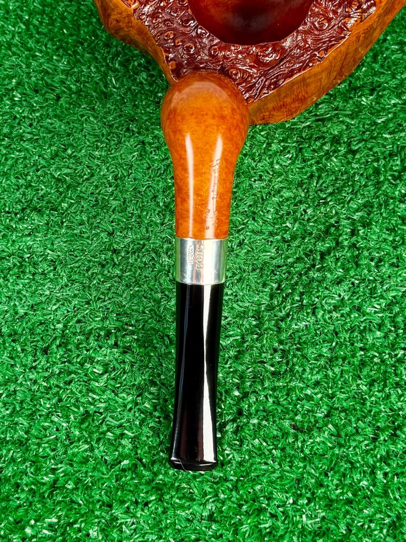 Puede incluir: Una pipa de tabaco con un hornillo de madera de brezo marr&oacute;n pulido con acabado texturizado, unido a un ca&ntilde;o de madera de tono m&aacute;s claro. Una banda de metal plateado separa el ca&ntilde;o de una boquilla c&oacute;nica negra, lisa y brillante.