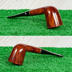 May include: A tobacco pipe featuring a polished brown wooden bowl with a visible grain pattern and a long, smooth black stem. The stem includes a small white circular inlay detail near the junction with the wooden bowl, creating a contrast.