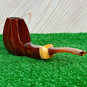 Pode incluir: Um cachimbo de tabaco com fornilho de madeira castanho-escura polida com um gr&atilde;o vertical liso. O cano liga-se a uma pe&ccedil;a de destaque em madeira natural de cor clara, que transita para uma piteira curva de acr&iacute;lico cor de &acirc;mbar.