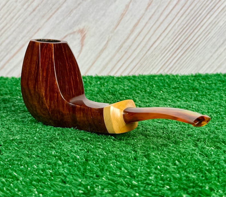 Pode incluir: Um cachimbo de tabaco com fornilho de madeira castanho-escura polida com um gr&atilde;o vertical liso. O cano liga-se a uma pe&ccedil;a de destaque em madeira natural de cor clara, que transita para uma piteira curva de acr&iacute;lico cor de &acirc;mbar.
