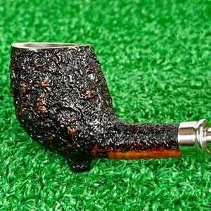 May include: A close-up of a tobacco pipe with a dark, textured bowl and a curved stem. The pipe has a silver band and a smooth, polished mouthpiece. The pipe is set against a green background.