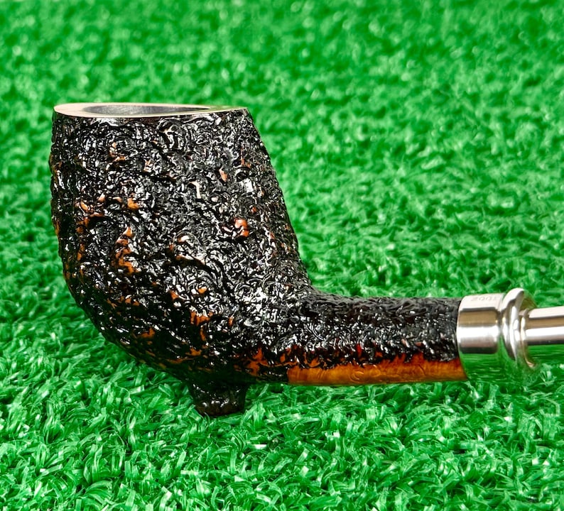 May include: A close-up of a tobacco pipe with a dark, textured bowl and a curved stem. The pipe has a silver band and a smooth, polished mouthpiece. The pipe is set against a green background.