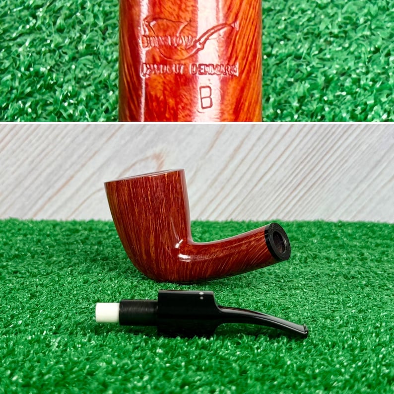 May include: A polished brown wooden tobacco pipe with a visible grain pattern. The pipe features a stamp reading "Stanwell Handout Denmark B". A separate black stem with a white filter tip is positioned in front of the pipe bowl on a green surface.