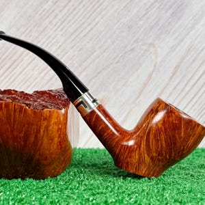 May include: A tobacco pipe with a polished brown wooden bowl featuring a prominent grain pattern, a silver-colored metal band, and a curved black acrylic stem. The pipe is positioned next to a large, rough-cut piece of matching brown wood.