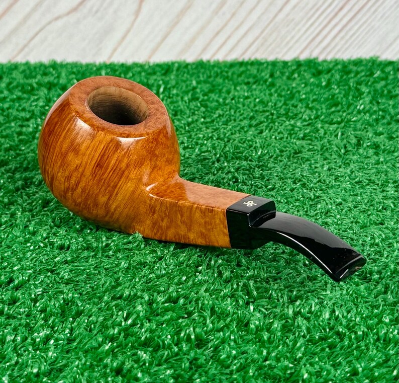 May include: A tobacco pipe with a polished light brown wooden bowl featuring a visible grain pattern and a smooth, curved black acrylic stem. The stem includes a small white logo detail near the junction where it connects to the wooden bowl.