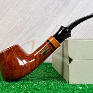 Pode incluir: Um cachimbo de tabaco castanho polido com um bocal preto. O cachimbo tem um design curvo com uma faixa castanha clara e uma pequena etiqueta dourada. O cachimbo repousa sobre uma superfície verde junto a uma caixa verde clara.