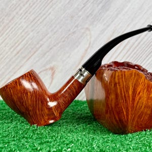 May include: A polished brown wooden tobacco pipe with a smooth, curved grain pattern, featuring a black acrylic stem and a silver-toned metal band connecting the bowl to the stem, resting against a large, textured block of matching brown wood.